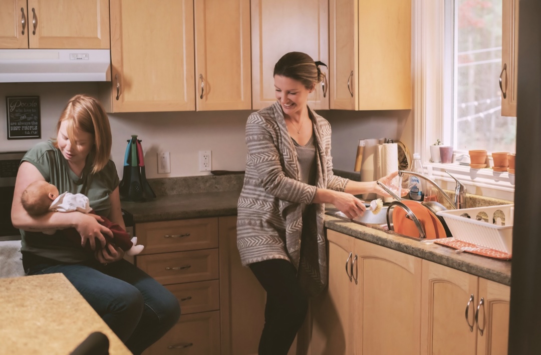 Doula standing in kitchen doing dishes and talking to a parent who is holding their baby.